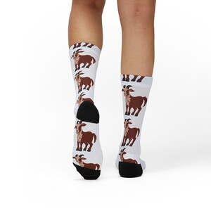 Puede incluir: Calcetines blancos con un patrón repetido de cabras marrones. Las cabras de dibujos animados tienen cuerpos marrones, caras blancas y cuernos curvos. Los calcetines tienen talones y dedos negros.