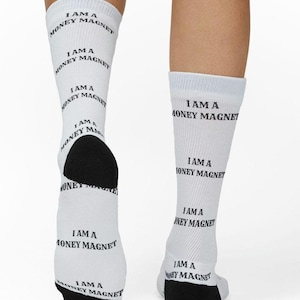 May include: A pair of white crew socks featuring the repeated black text "I AM A MONEY MAGNET" printed vertically along the leg and foot. The socks have solid black heels and toe sections, providing a distinct contrast to the white fabric.