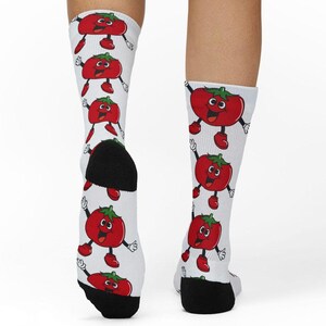 May include: White crew socks featuring a repeating pattern of cartoon tomatoes with green stems, smiling faces, black arms, and red shoes. The socks have solid black heels and toes, providing a distinct contrast to the white fabric and red tomato print.
