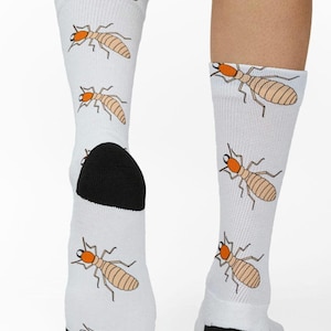 Könnte beinhalten: Weiße Socken mit einem sich wiederholenden Muster aus Cartoon-Termiten in Beige und Orange. Die Socken haben schwarze Fersen und Zehen. Ein lustiges und skurriles Accessoire.