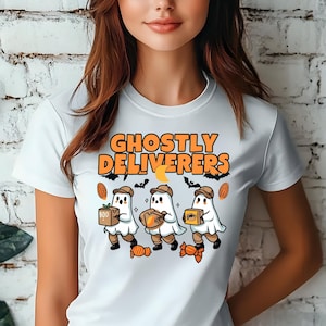 May include: White t-shirt featuring the text "Ghostly Deliverers" in orange, with illustrations of three ghost characters carrying boxes and candy. The design includes bats, a crescent moon, and candy corn.