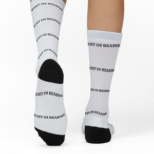 Op de afbeelding: Een paar witte sokken met de herhaalde zwarte tekst "QUIET IM READING" horizontaal op de stof gedrukt. De sokken hebben effen zwarte hielen en tenen, wat een duidelijk contrast vormt met de witte basis van het kledingstuk.