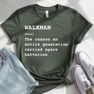 Pode incluir: Uma t-shirt de manga curta verde-oliva com texto branco que diz: WALKMAN [Noun] The reason an entire generation carried spare batteries. A peça tem gola redonda e mangas dobradas, apresentada com calções de ganga azul-claros e sapatilhas brancas.