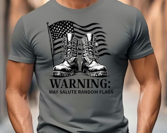 Może przedstawiać: Szary t-shirt z krótkim rękawem z czarno-białą grafiką przedstawiającą dwa buty wojskowe na tle amerykańskiej flagi. Pod butami znajduje się napis: WARNING: MAY SALUTE RANDOM FLAGS. Wzór jest wyśrodkowany na klatce piersiowej koszulki.