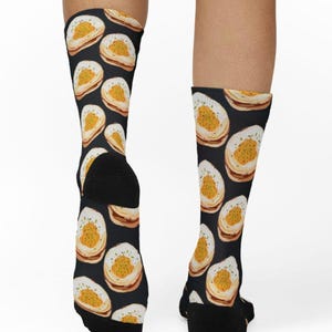 Peut inclure: Chaussettes noires avec un motif répété d'un aliment ressemblant à un œuf au plat. Le motif en forme d'œuf a un anneau extérieur blanc, un centre jaune et des herbes vertes saupoudrées. Les chaussettes sont portées sur les jambes.