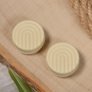 May include: Two off-white round ceramic coasters with a raised geometric design. The coasters are on a wooden surface.
