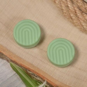 May include: Two green round silicone buttons with a raised geometric pattern. The buttons are on a wooden surface.