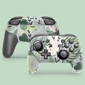 Peut inclure: Deux manettes Nintendo Switch Joy-Con avec un motif floral vert et blanc. Les manettes sont noires et ont des boutons marqués A, B, X, Y et un signe plus.