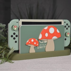 May include: A mint green Nintendo Switch console with mushroom-themed accessories. The console has a green base, a green card holder with two mushroom cutouts, and mushroom-shaped thumb grips. The mushrooms have red caps with white spots.