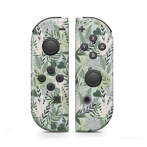 Puede incluir: Un par de mandos Nintendo Switch Joy-Con verdes y blancos con un patrón de hojas. Los mandos tienen botones negros y un botón de inicio.