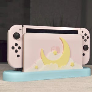 Peut inclure: Un support de console Nintendo Switch rose et bleu avec un motif de lune et de nuage. Le support a une découpe pour la console et un emplacement pour les Joy-Cons.