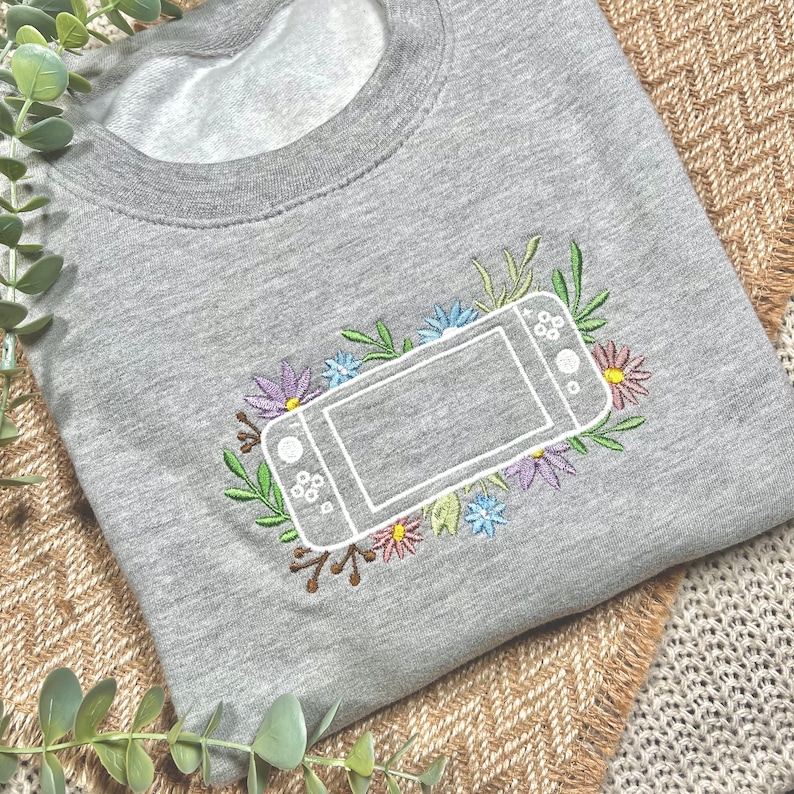 以下が含まれることがあります： ゲーム機のアウトラインが白で刺繍された、グレーのトレーナー。カラフルな花と緑の葉で囲まれています。長袖トップの胸の中央にデザインがあります。