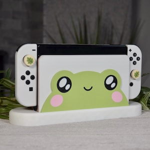 May include: White Nintendo Switch console stand with a green frog design. The stand has a white base and a green frog face with black eyes and pink cheeks. The console is white with black buttons and green leaf-shaped buttons.
