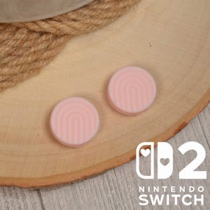 以下が含まれることがあります： 虹のデザインが施された、ピンク色のNintendo Switch用サムグリップが2つ。丸いグリップが木製の表面に置かれています。Nintendo Switchのロゴが右下に表示されています。