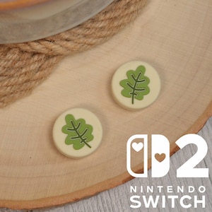 以下が含まれることがあります： 緑の葉のデザインが施された、クリーム色のNintendo Switch用サムグリップが2つ。木製の表面に配置され、Nintendo Switchのロゴとテキストが白で表示されています。グリップは小さく丸いです。