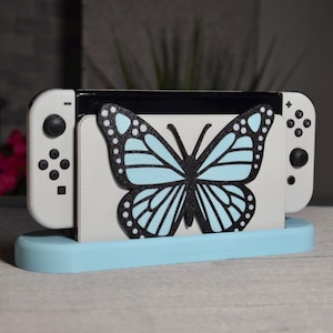 May include: A light blue stand with a butterfly design holds a white Nintendo Switch console. The console is in a closed position with the Joy-Con controllers attached.