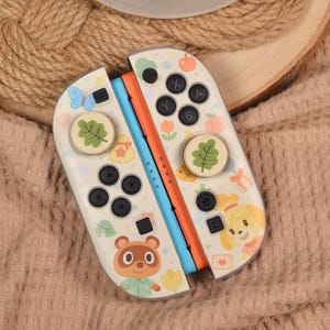 May include: White Nintendo Switch Joy-Con controllers with colourful illustrations. The controllers feature images of a butterfly, leaves, fruit, and cartoon characters. The buttons are black, and the sides are blue and orange.