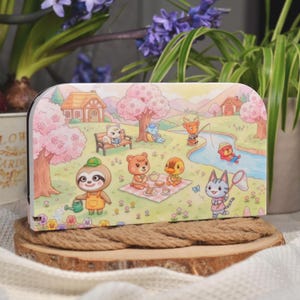 Può includere: Una custodia rettangolare bianca con un'illustrazione colorata di animali dei cartoni animati in un parco. La scena include case, alberi, un fiume e vari animali. La custodia è esposta su una superficie di legno.