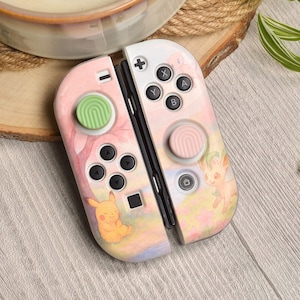 Puede incluir: Mandos Joy-Con de Nintendo Switch rosas y blancos con un diseño de acuarela con Pikachu y otros personajes. Los mandos tienen botones negros y joysticks verdes y rosas.