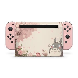 以下が含まれることがあります： ピンク色のJoy-Conコントローラーを備えたNintendo Switchコンソール。コンソールは、桜のデザインと漫画のキャラクターを備えたカスタムスキンを備えています。スキンは、ピンクの花と灰色のキャラクターを備えた淡いベージュの背景を持っています。