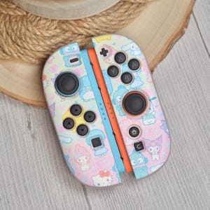 Peut inclure: Une manette Joy-Con Nintendo Switch colorée avec un motif pastel représentant des personnages de dessins animés. La manette a des boutons et des joysticks noirs, avec un centre bleu et orange. Le design comprend Hello Kitty et d'autres personnages mignons.