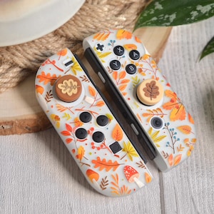 Może przedstawiać: Para białych kontrolerów Nintendo Switch Joy-Con z jesiennym wzorem. Kontrolery mają białe tło z pomarańczowymi, żółtymi i brązowymi liśćmi, grzybami i jagodami. Kontrolery mają również brązowe przyciski w kształcie liści.