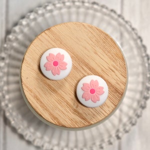 以下が含まれることがあります： ピンクの桜の花柄の白いシリコン製携帯電話グリップ2個。