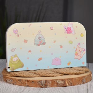 May include: A rectangular console cover featuring a pastel gradient background transitioning from pale yellow to light blue. The surface is decorated with various small cartoon characters, including a pink creature, a green figure, and a yellow mouse.