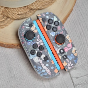 以下が含まれることがあります： ピンク、青、オレンジの花柄が施されたグレーのNintendo Switch Joy-Conコントローラー。 コントローラーには黒いボタンとジョイスティックがあり、オレンジと青のアクセントが付いています。 デザインは、さまざまな花と葉が特徴です。