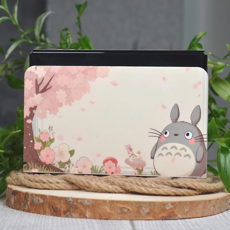 May include: A white and pink Nintendo Switch case with a cartoon image of Totoro from the movie My Neighbor Totoro. The case features a pink cherry blossom tree and pink petals falling from the tree.