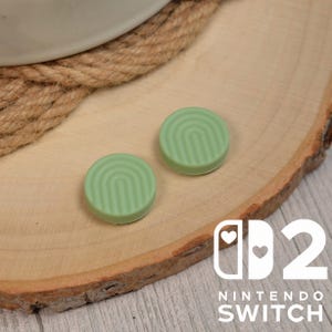 以下が含まれることがあります： 溝のある虹のデザインが施された、2 つの淡い緑色の Nintendo Switch ジョイスティックキャップ。キャップは、白い Nintendo Switch ロゴが入った木製の表面にあります。