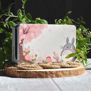 May include: White Nintendo Switch console skin with a pink cherry blossom tree and a grey Totoro character from the movie My Neighbor Totoro. The skin is on a wooden base with a rope around it.