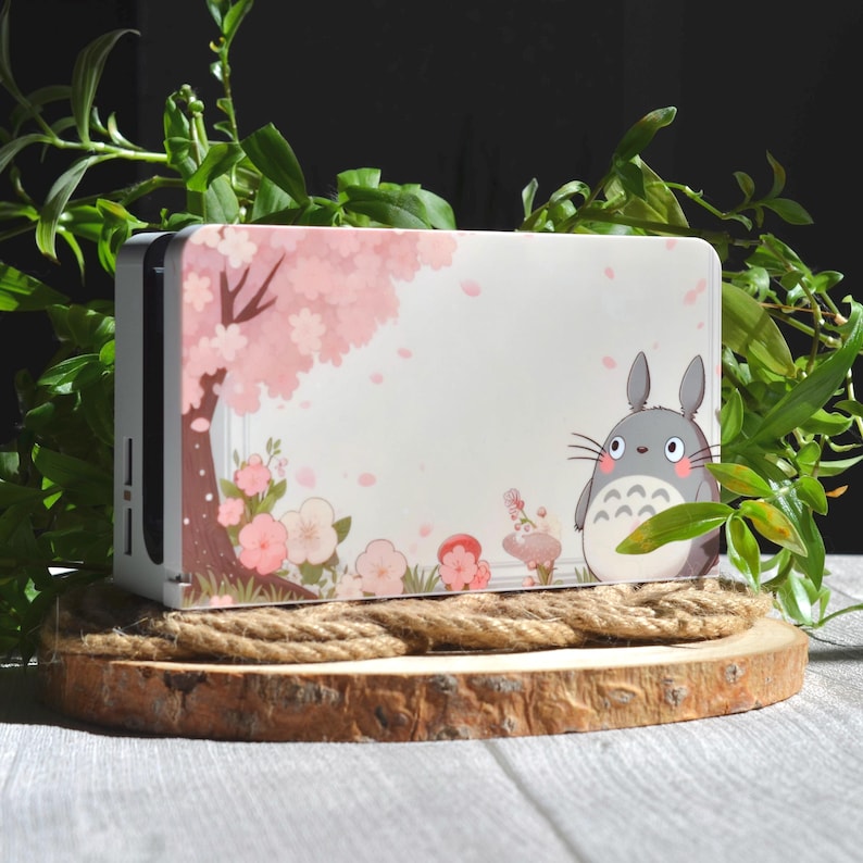 May include: White Nintendo Switch console skin with a pink cherry blossom tree and a grey Totoro character from the movie My Neighbor Totoro. The skin is on a wooden base with a rope around it.