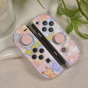 Puede incluir: Un mando de Nintendo Switch rosa y blanco con un diseño de temática Pokémon. El mando tiene un Pikachu, un Eevee y otros personajes de Pokémon. El mando tiene botones rosas y un diseño rosa y blanco.