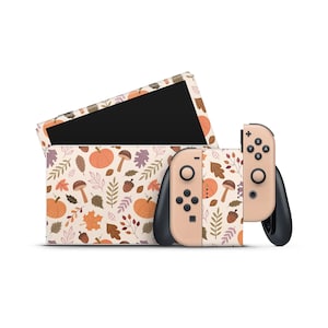 Puede incluir: Un conjunto de consola Nintendo Switch y mandos Joy-Con con un diseño de temática otoñal. El diseño presenta un patrón de calabazas naranjas, setas marrones, hojas rojas y naranjas y bellotas marrones sobre un fondo crema.