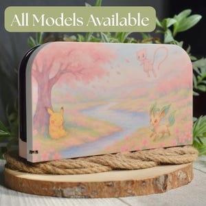 以下が含まれることがあります： ピンクと白のNintendo Switchドック。桜の風景の中にピカチュウ、ミュウ、リーフィアが描かれたイラストが特徴です。「All Models Available」の文字が上部にあります。