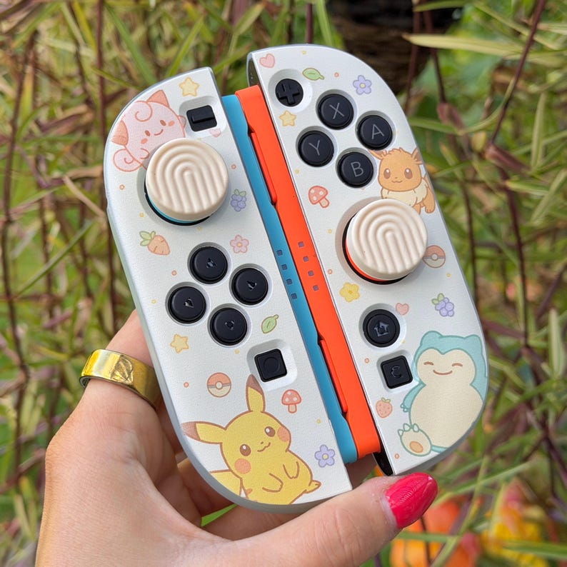 May include: A pair of white game controllers featuring colorful illustrations of Pikachu, Eevee, Snorlax, and Jigglypuff. The controllers include black buttons, light beige thumbstick covers with a ridged pattern, and small icons of flowers, mushrooms, and berries.