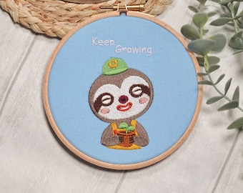May include: Embroidered hoop art featuring a sloth wearing a green hat and holding a potted plant. The background is light blue and the text "Keep Growing." is embroidered in white.