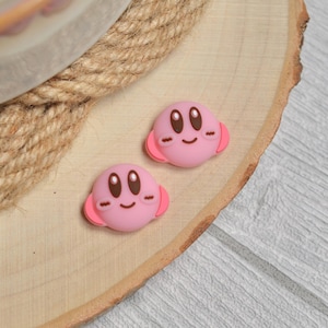 May include: Two pink cartoon character charms with brown eyes and smiling faces. The charms have pink ear-like features on the sides. They are placed on a wooden surface.