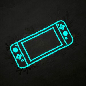 以下が含まれることがあります： 暗い背景に、光るターコイズ色の携帯ゲーム機の輪郭。コンソールには画面、ボタン、コントロールスティックがあります。画像は柔らかく、テクスチャード加工された外観で、デジタルイラストまたは様式化されたデザインを示唆しています。