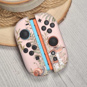 Peut inclure: Manettes Joy-Con Nintendo Switch roses avec un motif sur le thème de la nature. Les manettes présentent une illustration de fougère, de champignon et d'escargot. Le contrôleur gauche a un accent bleu et le droit un accent orange.