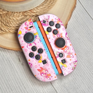 May include: Pink Nintendo Switch Joy-Con controllers with a cartoon character design and colourful stars. The controllers have black buttons and joysticks, with orange and blue accents.