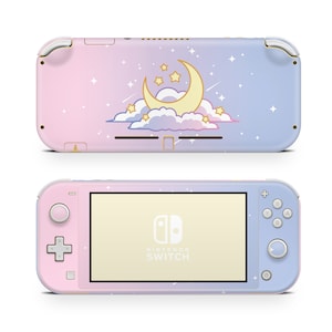 Puede incluir: Una consola Nintendo Switch Lite rosa y azul con un diseño de luna y estrellas. La consola tiene una pantalla blanca y el logotipo de Nintendo Switch es visible en la parte delantera.