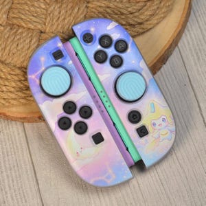 May include: A pair of game controllers featuring a pastel sky design with clouds, stars, and constellations. The controllers have light blue thumbstick covers, black buttons, and a graphic of a smiling moon and a yellow star-shaped creature.