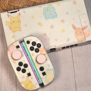 May include: A gaming console and two controllers featuring a cream-colored skin with pastel illustrations of various creatures, including Pikachu, Bulbasaur, Eevee, and Snorlax, alongside small icons of flowers, stars, hearts, and berries.