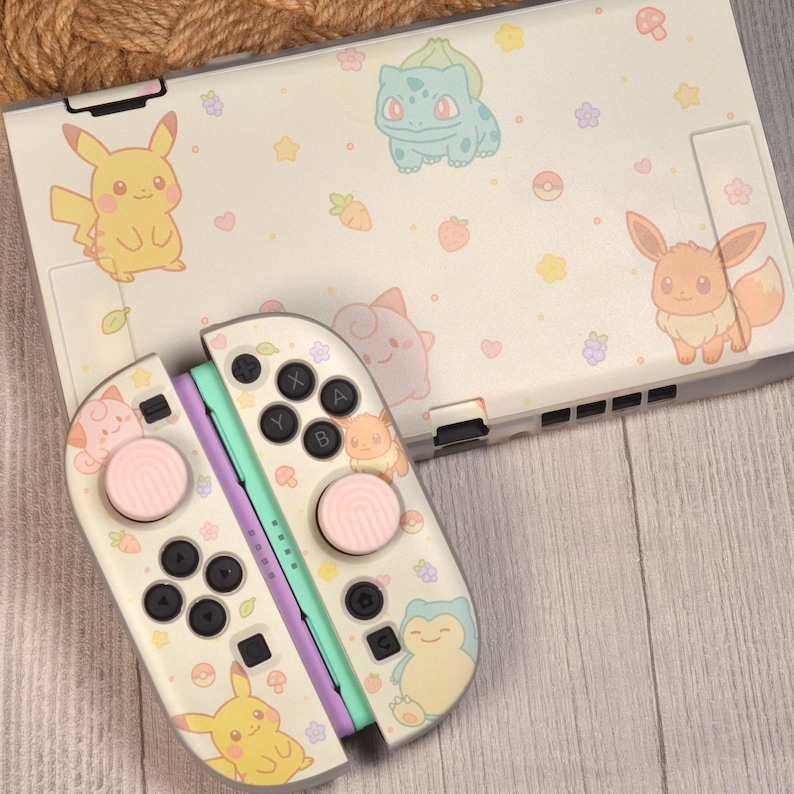May include: A gaming console and two controllers featuring a cream-colored skin with pastel illustrations of various creatures, including Pikachu, Bulbasaur, Eevee, and Snorlax, alongside small icons of flowers, stars, hearts, and berries.