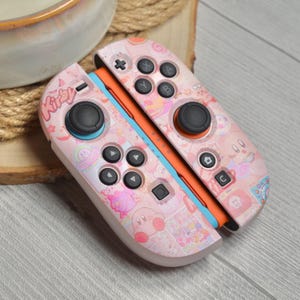 Pode incluir: Controladores Joy-Con Nintendo Switch rosa e brancos com um design Kirby. Os controladores têm botões e joysticks pretos, com detalhes em laranja e azul. A palavra "Kirby" está impressa no controlador esquerdo.