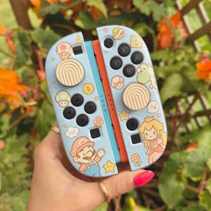 May include: A light blue Nintendo Switch Joy-Con controller with cartoon graphics of Mario, Princess Peach, and other characters. The controller has black buttons, beige joysticks, and orange and blue accents.
