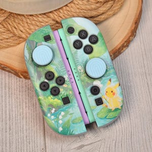 May include: A pair of game controllers featuring a light green and blue forest-themed design with lily of the valley flowers. A yellow Pikachu character wearing a raincoat is printed on the right side. Both controllers have light blue thumbstick covers.