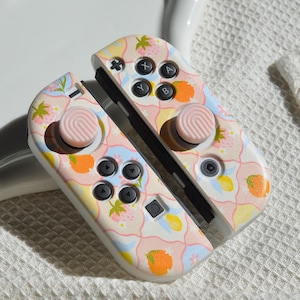 Puede incluir: Un mando Nintendo Switch Joy-Con rosa y blanco con un patrón floral y detalles en naranja. El mando tiene botones rosas y un diseño rosa y blanco en la parte posterior.
